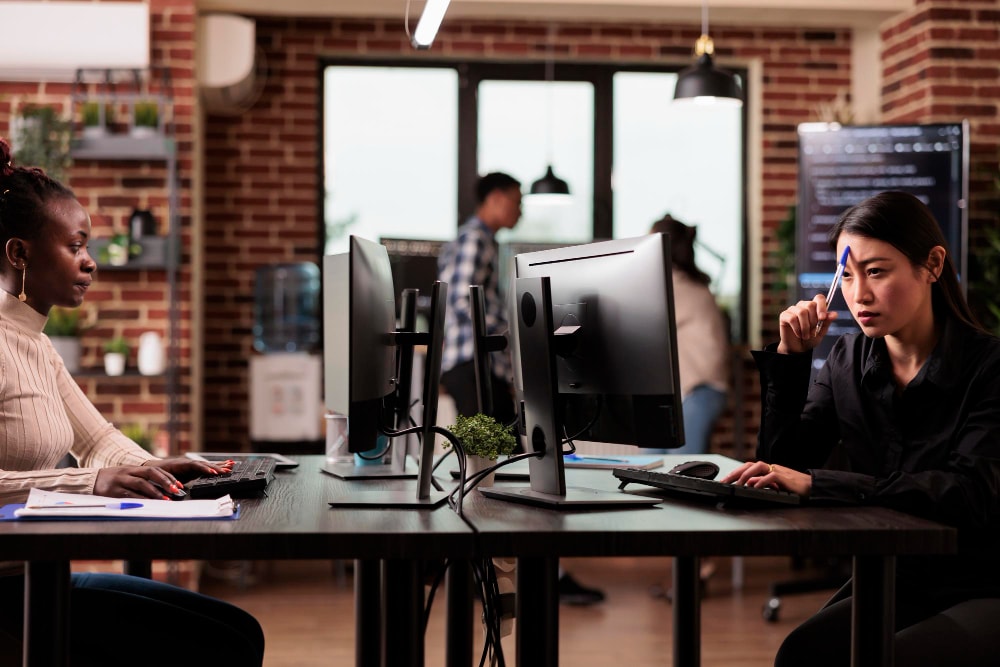 A group of four customer service agents wearing headsets work at their computers, smiling and communicating in a high-rise office with large windows.