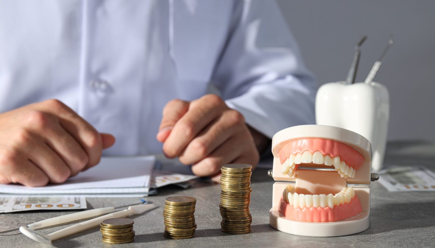 Tannlegepriser i Oslo: Hva bør du vite? 11 Tannlege ved skrivebord med kunstige tenner og stabler med mynter som symboliserer tannlegekostnader.