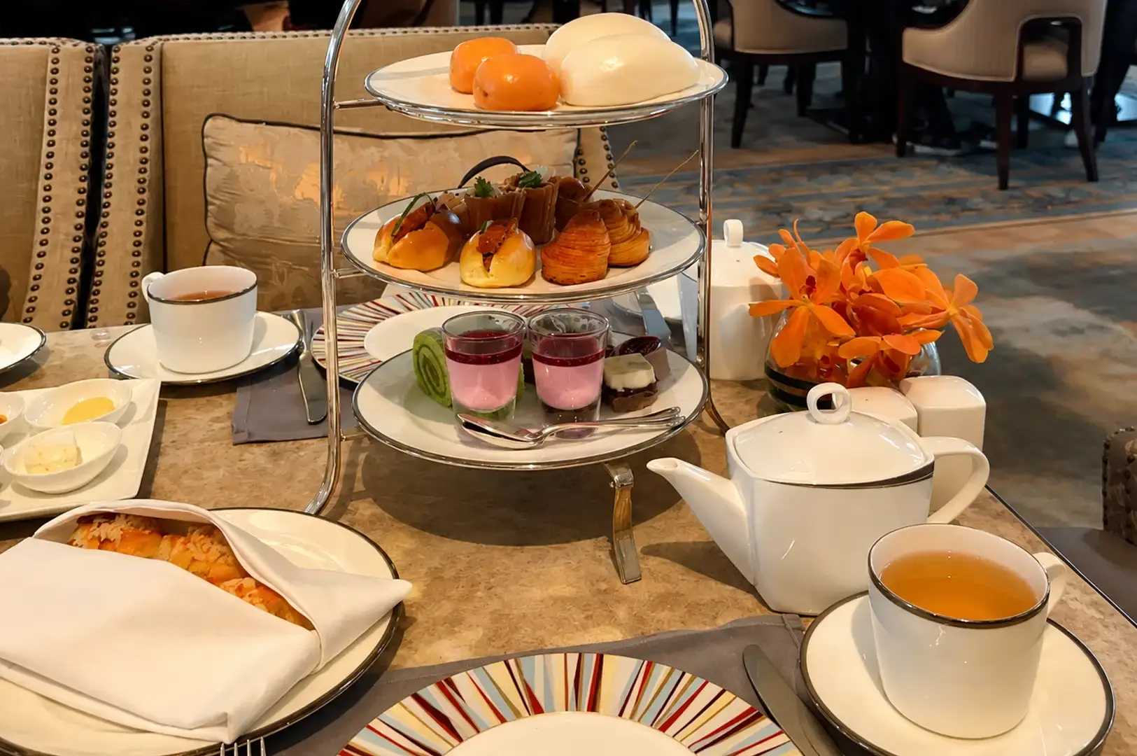 A full afternoon tea table layout featuring a three-tiered stand filled with savory pastries, bao buns, and dessert shooter glasses, flanked by a white ceramic teapot, teacups with saucers, a vase of vibrant orange orchids, and warm baked scones wrapped in a white cloth napkin on a plate.