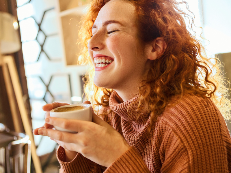 Mulher ruiva toma café e sorri. Imagem: Nastasic de Getty Images Signature -  Canva.