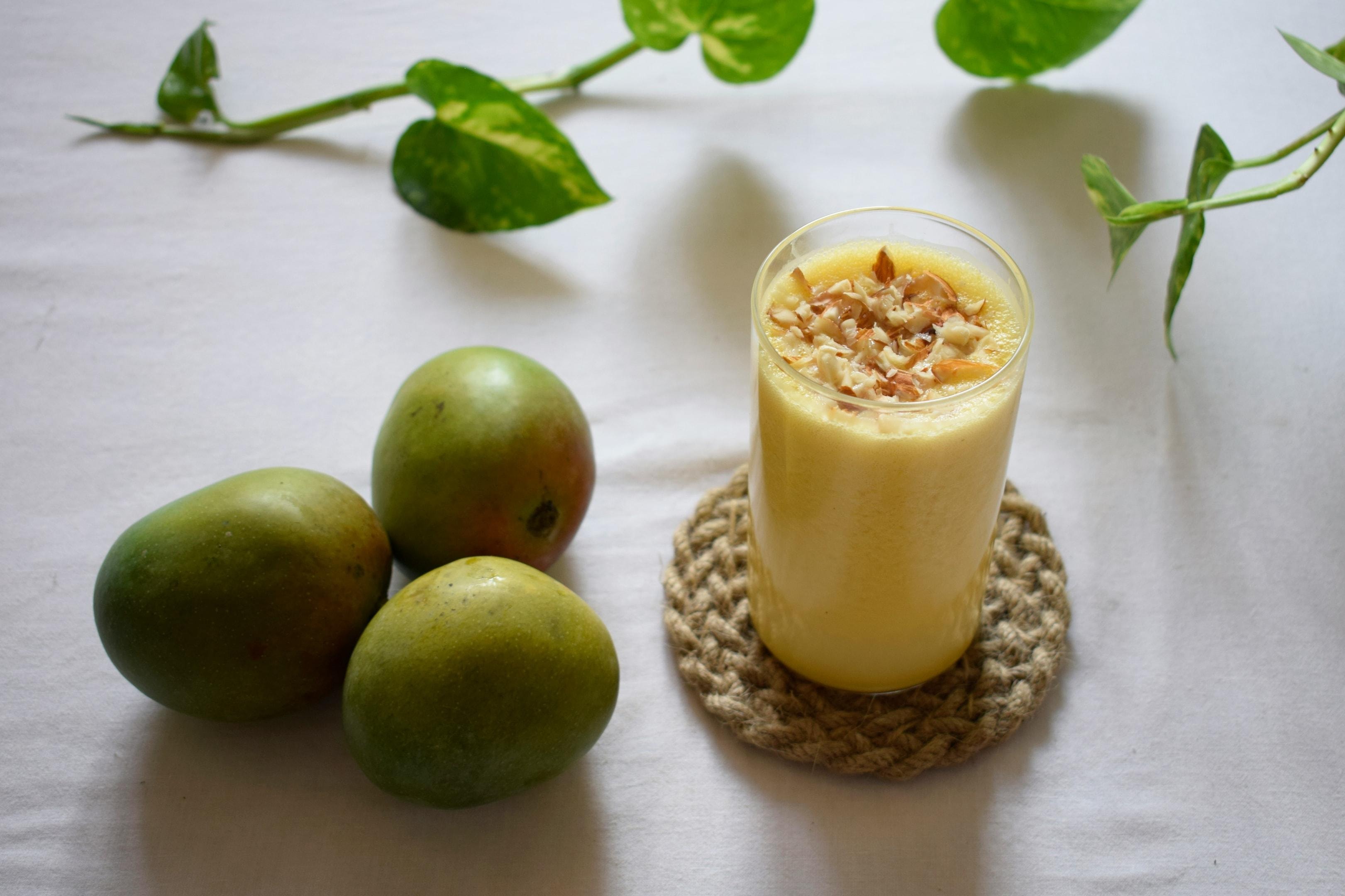  This image features a tall glass of creamy yellow mango lassi topped with sliced nuts, resting on a woven coaster. To the left, three whole green-skinned mangoes sit on a white surface, while a green vine with heart-shaped leaves stretches across the background.
