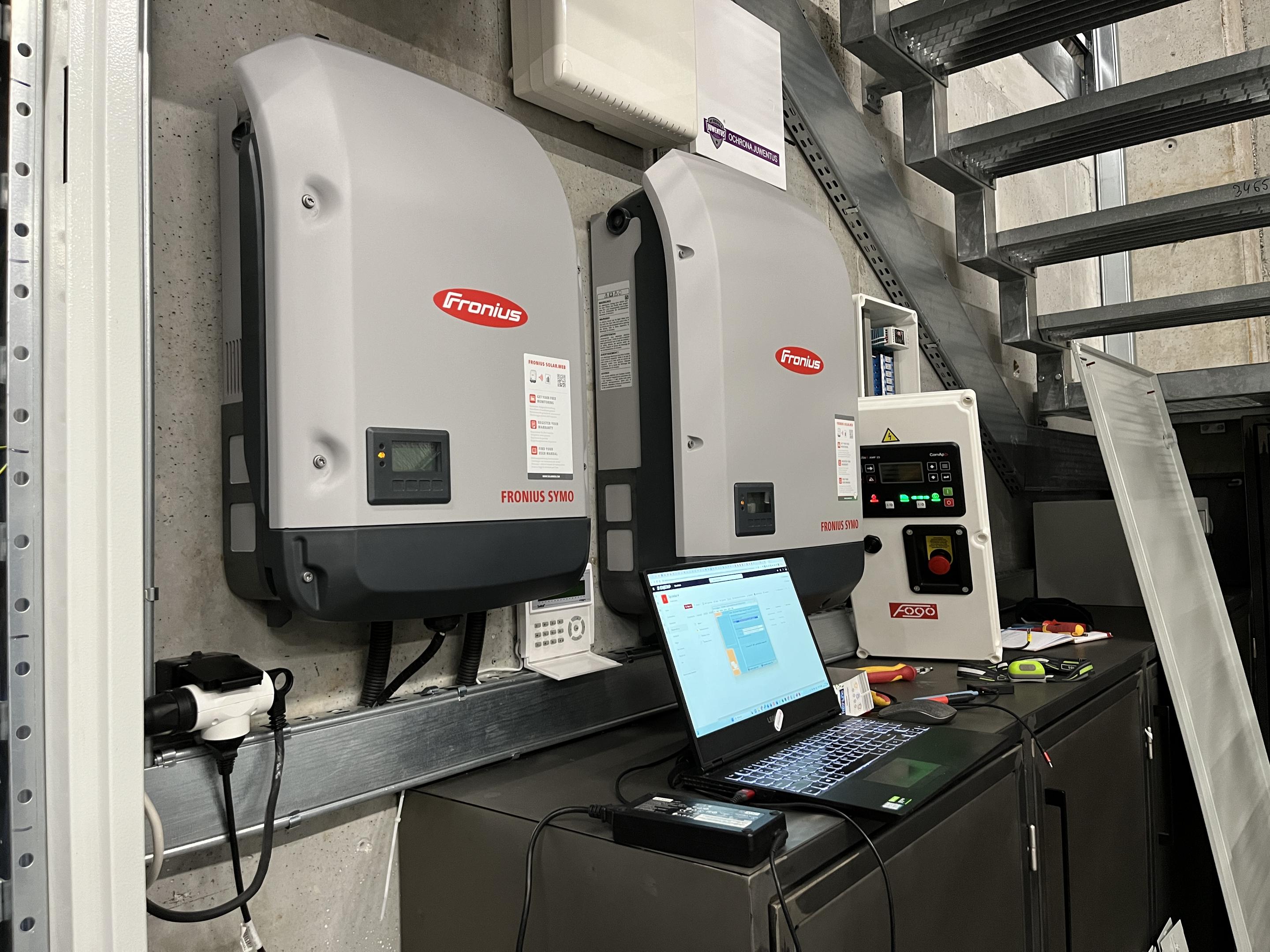 Na zdjęciu widać instalatora sprawdzającego rozdzielnicę elektryczną w jasnym pomieszczeniu technicznym. Mężczyzna zajmuje się efektywnym zarządzaniem energią, co może obejmować instalację fotowoltaiczną oraz magazynowanie energii dla zapewnienia niezawodności dostawy energii.