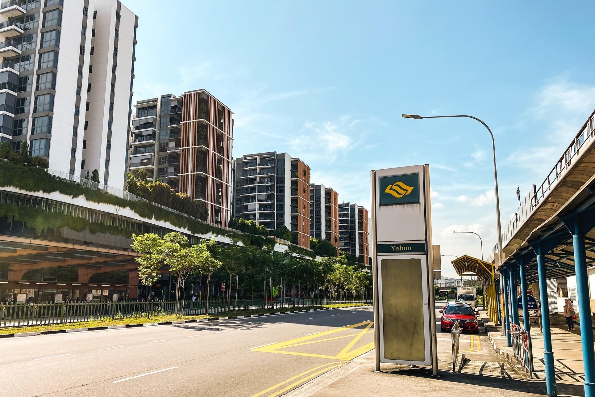  Modern residential towers with distinctive vertical fins and greenery-lined balconies stand along a wide, sunlit road. A prominent sign for Yishun is visible next to a taxi stand and sheltered walkway, under a clear blue sky.