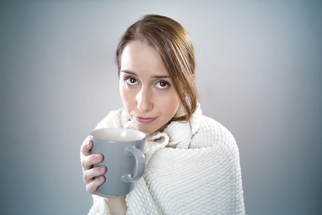 An image of a sick young woman with a blanket around her shoulders sipping soothing hot tea.