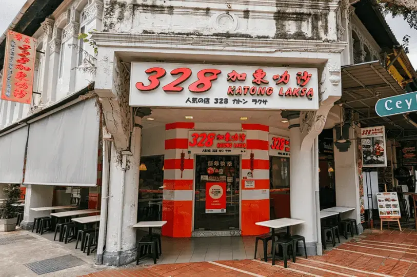 The exterior of the famous "328 Katong Laksa" restaurant located in a white colonial-style corner building. The storefront features a prominent white sign with red lettering and distinctive orange-and-white striped pillars. Outdoor seating with simple black stools and white tables lines the sidewalk.