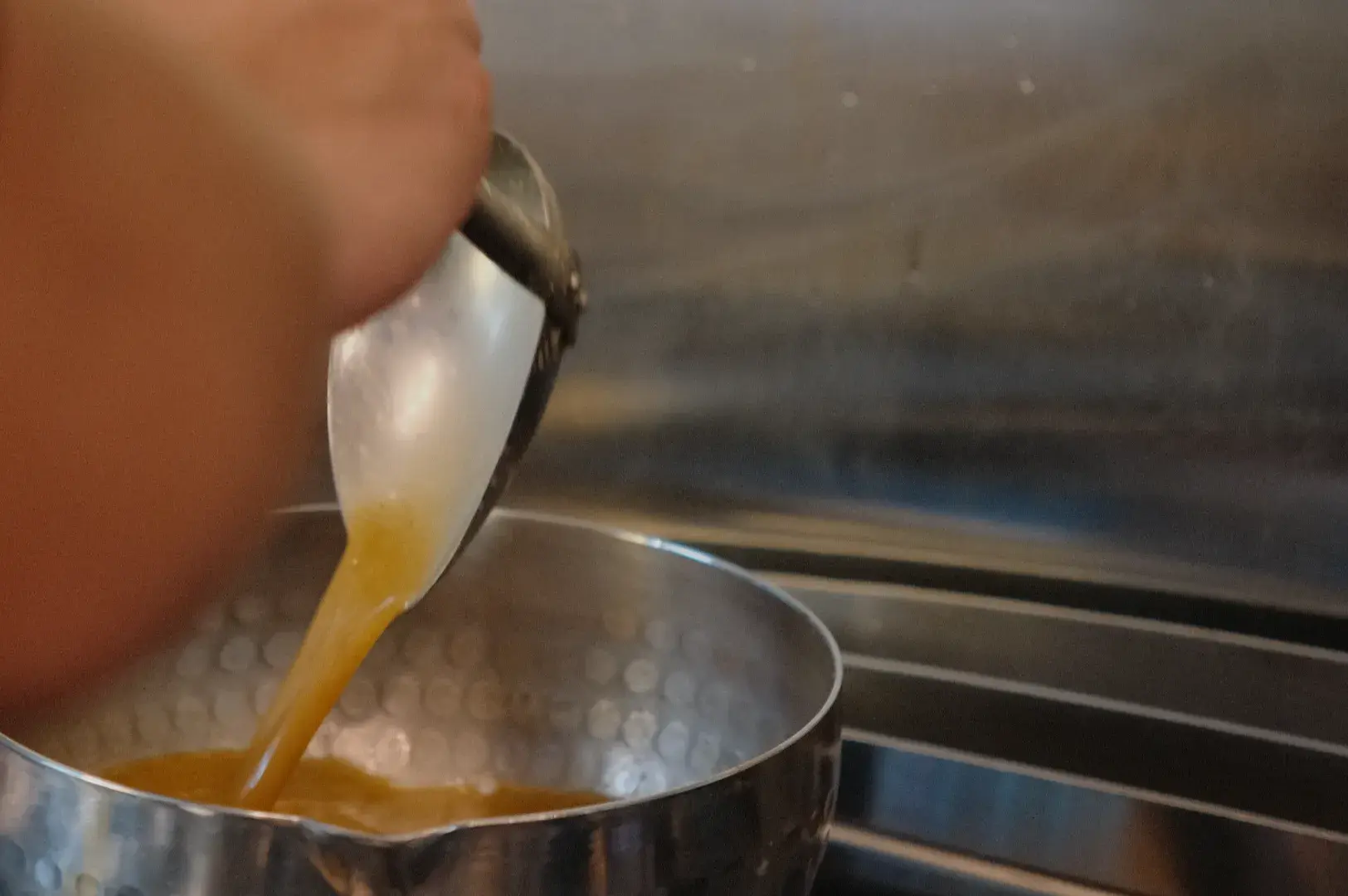 A hand pours a raw egg from a ladle into a stainless-steel saucepan on a stovetop, capturing a close-up moment of everyday cooking.