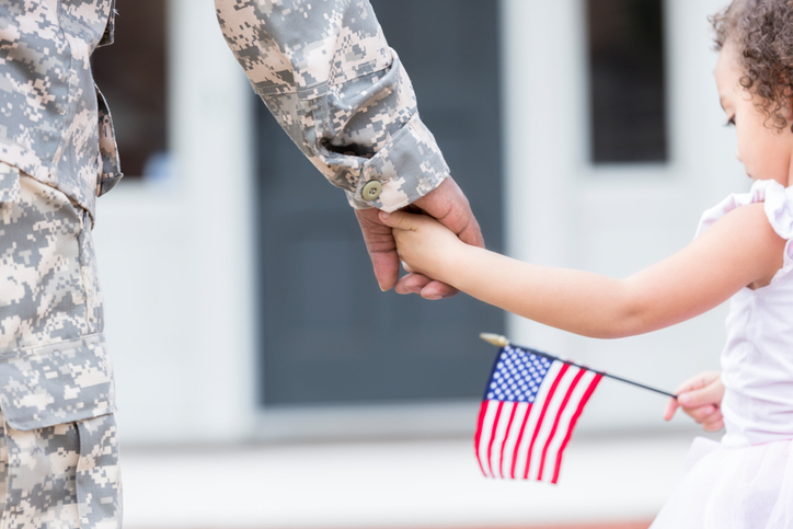 Mom in fatigues holding her little girl's hand as she holds a U.S. flag. 