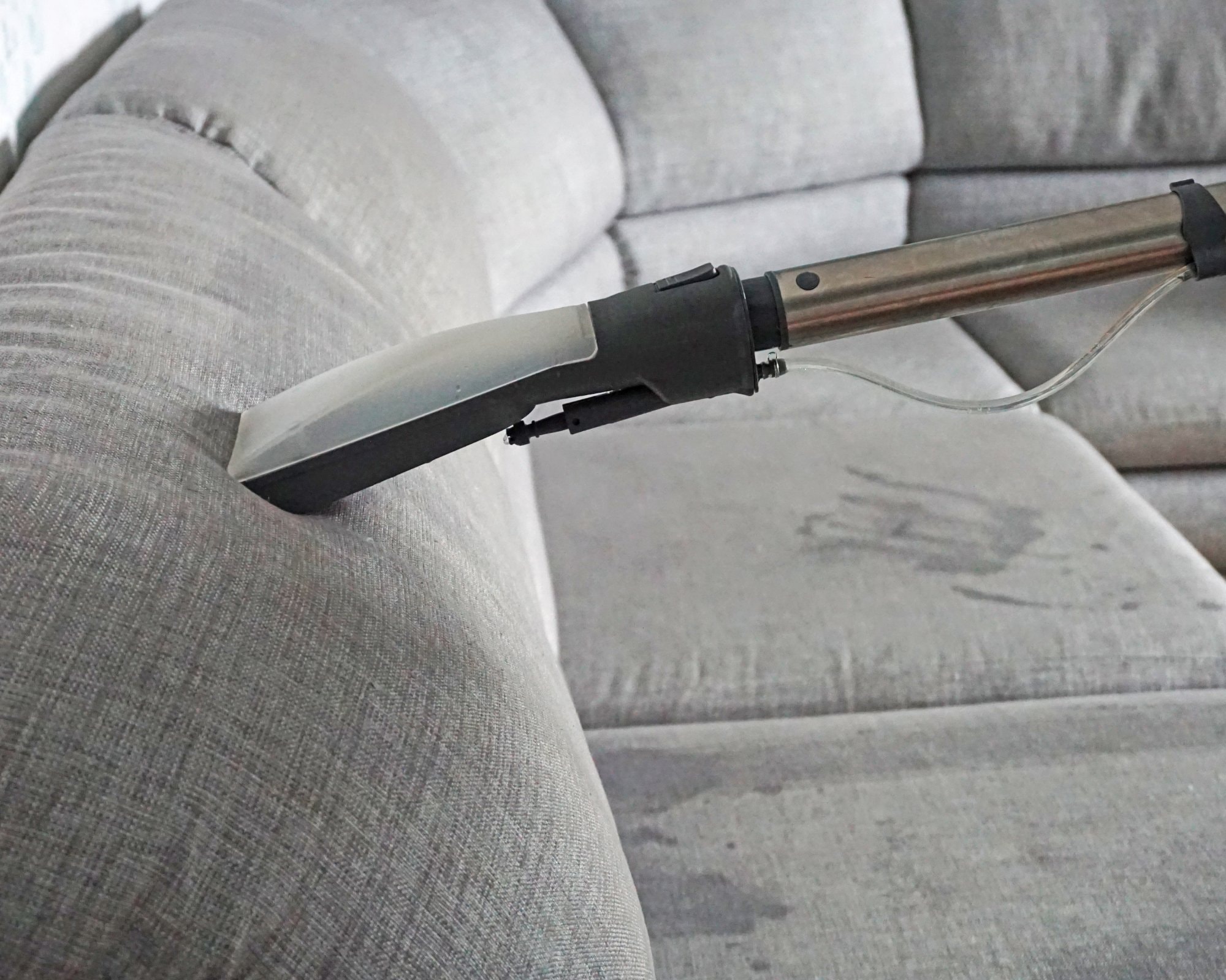 Close-up of a professional upholstery cleaning tool in use on a light gray fabric sectional sofa, with visible damp spots from deep cleaning.