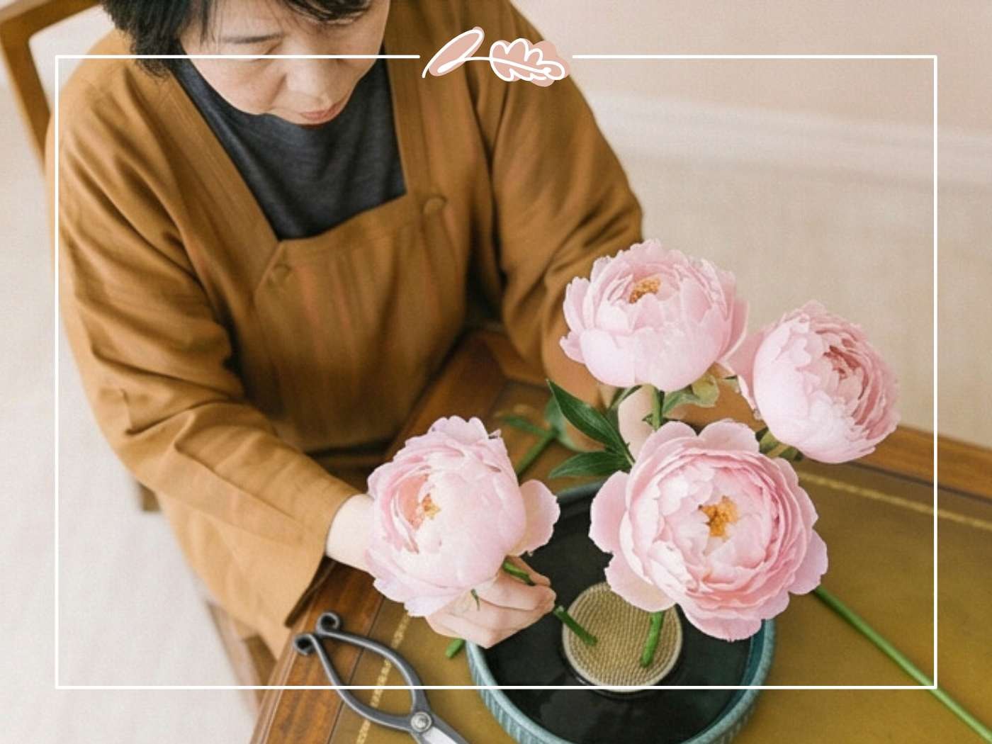 Woman in ochre linen jacket arranging pink peonies in a ribbed teal ceramic ikebana vessel on a wooden table | The Art of Ikebana | Fabulous Flowers & Gifts