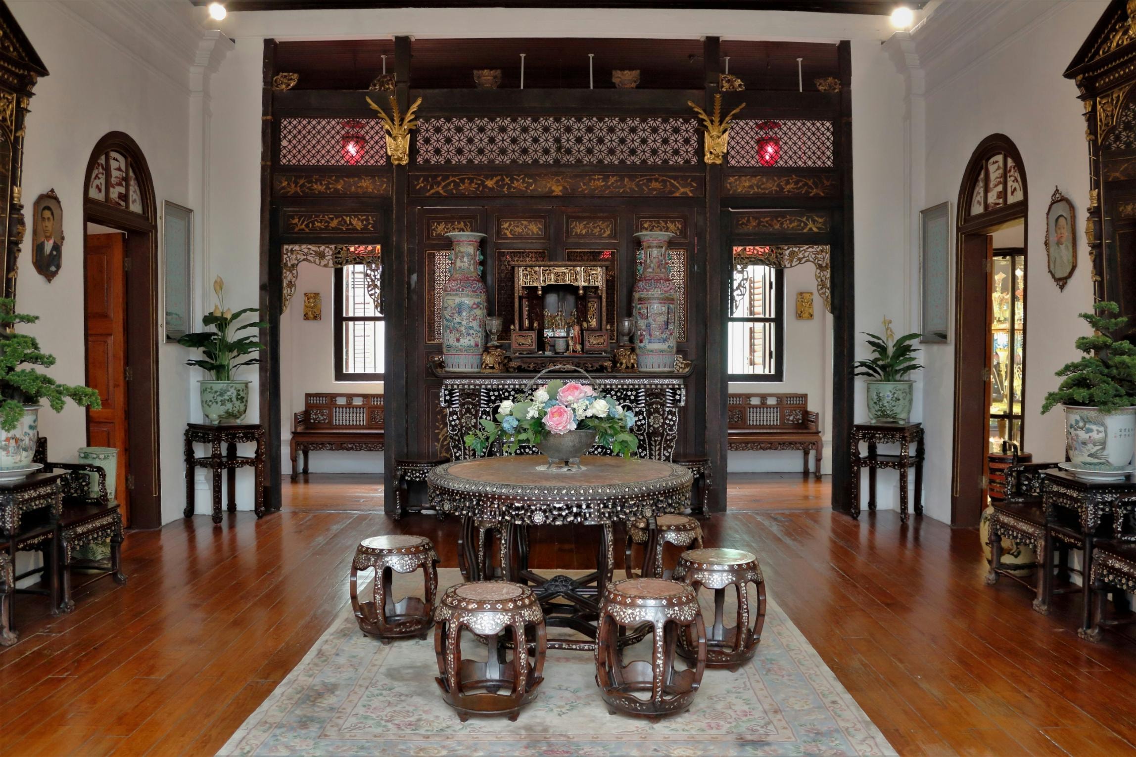  This interior view showcases a traditional Peranakan reception hall featuring dark, intricately carved wooden partitions and mother-of-pearl inlaid furniture. A circular table with matching stools sits on a decorative rug in the center of the room, flanked by ornate vases and potted plants that lead the eye toward an elaborate altar in the background.