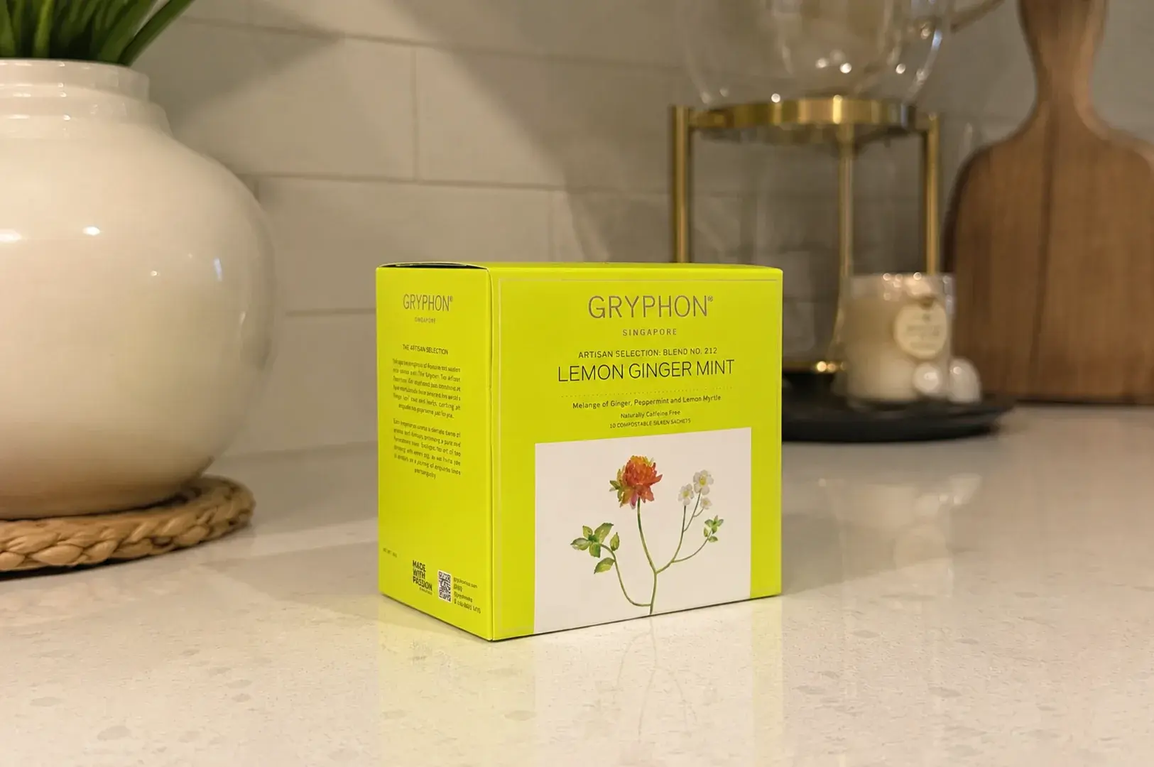 A chartreuse-yellow box of Gryphon Singapore's Artisan Selection Blend No. 212, "Lemon Ginger Mint," is positioned at an angle on a clean, polished white countertop. The front label shows a minimalist botanical drawing of a single red clover blossom alongside small white flowers, indicating a caffeine-free herbal tisane with peppermint, ginger, and lemon myrtle. The background features a crisp white tiled backsplash, a white ceramic vase resting on a woven coaster to the left, and a blurred wooden cutting board and a lit candle near a glass tea maker to the right.