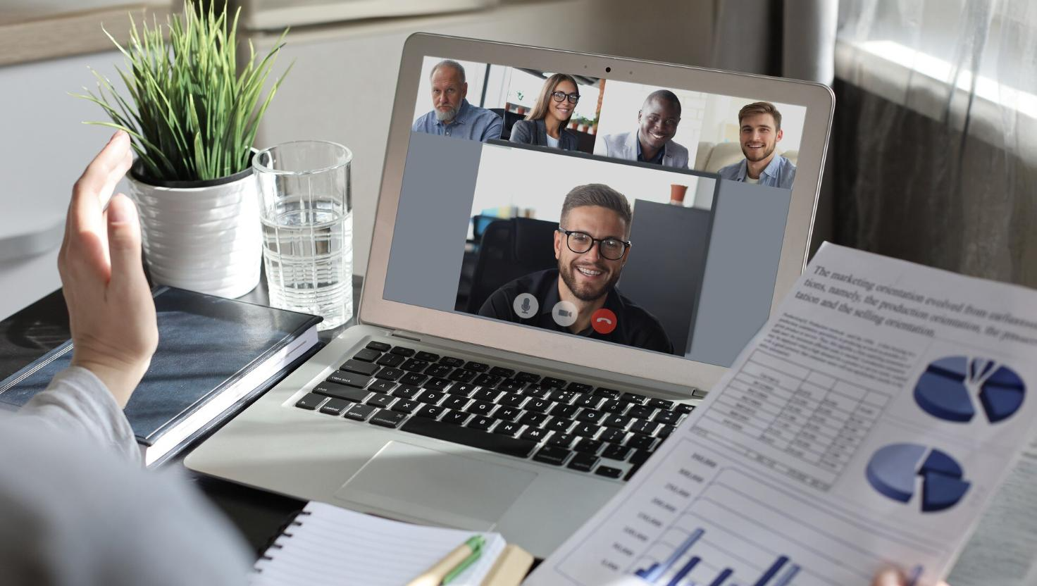 A person attends a virtual meeting on a laptop, with five diverse colleagues displayed in a video conferencing grid. A report with charts is held in one hand, symbolizing data-driven remote collaboration through business communication tools.
