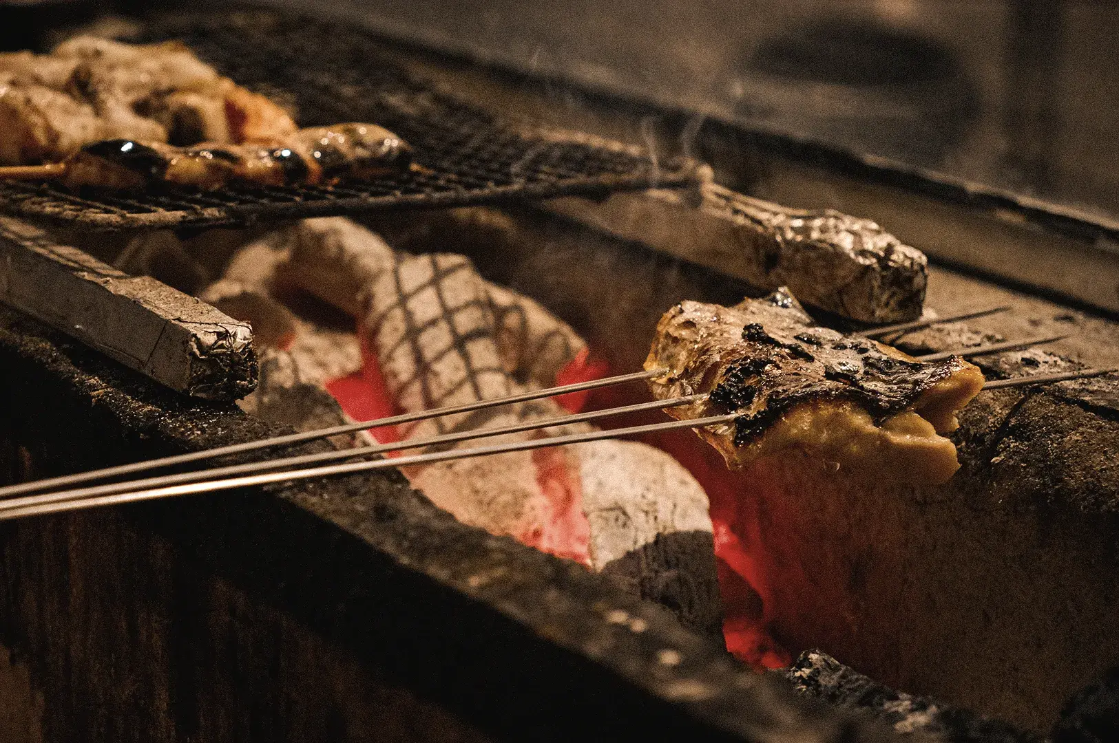A low-angle close-up shot showcases seafood skewers grilling over glowing red binchotan white charcoal on the Sumi grill counter at at Sushi to Sumi.