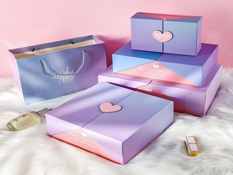 A picture of pastel-colored subscription boxes and a matching gift bag arranged on a soft white surface, featuring pink and purple tones with heart details.