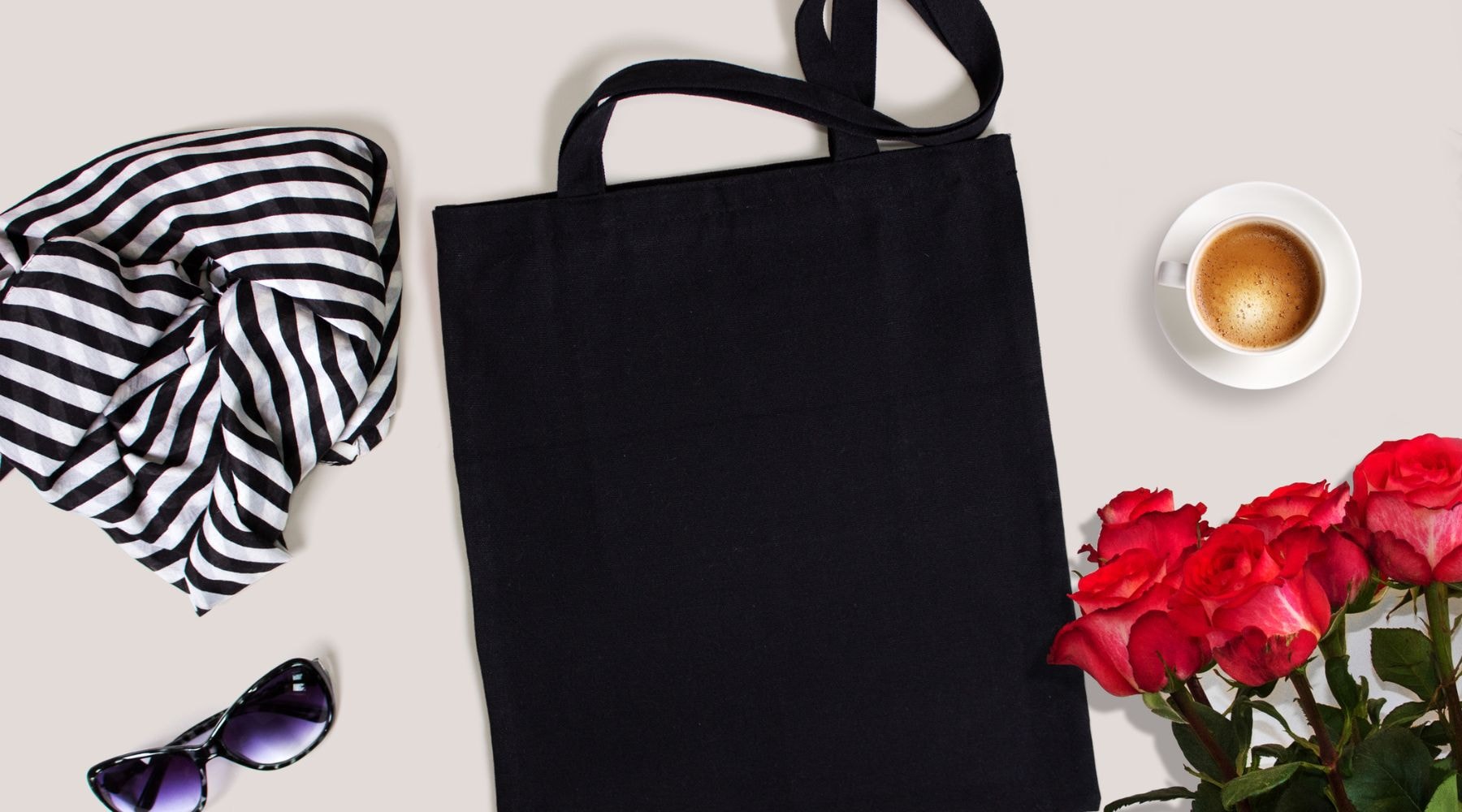 Black tote bag styled with striped scarf and sunglasses next to a cup of coffee and roses.