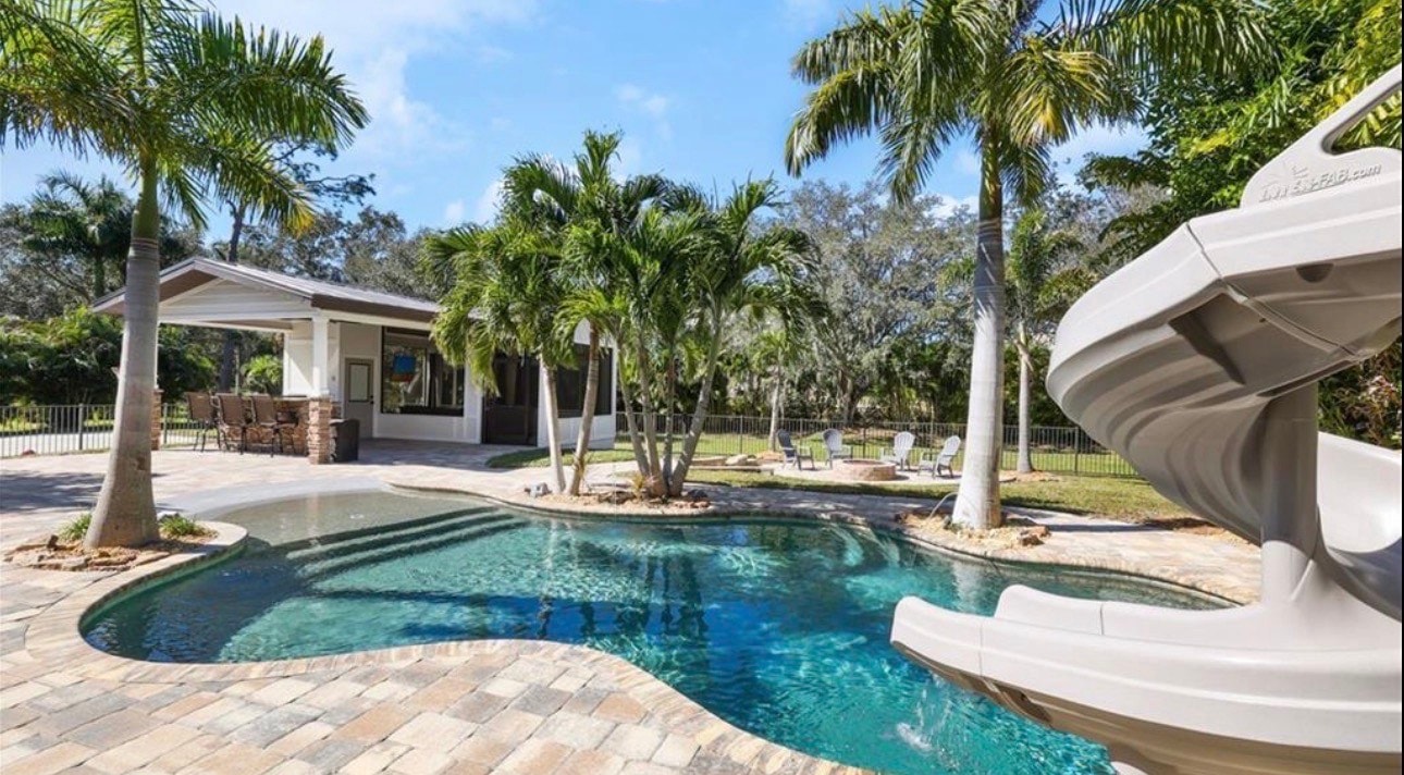 The image depicts a resort-style pool area featuring a spacious outdoor kitchen and entertaining space, all surrounded by lush landscaping, creating an inviting atmosphere for relaxation and gatherings in Livingston Woods, Naples. This expansive outdoor living space is perfect for enjoying Florida's beautiful weather.