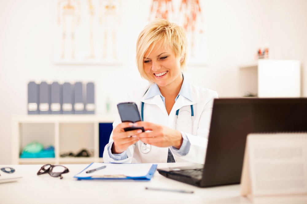 A cheerful female doctor with a short blonde haircut and white lab coat checks her smartphone while seated at her office desk. Unified Communications systems and mobile-friendly team collaboration tools contribute to seamless communication with patients and colleagues, supporting higher business productivity in clinical workflows.