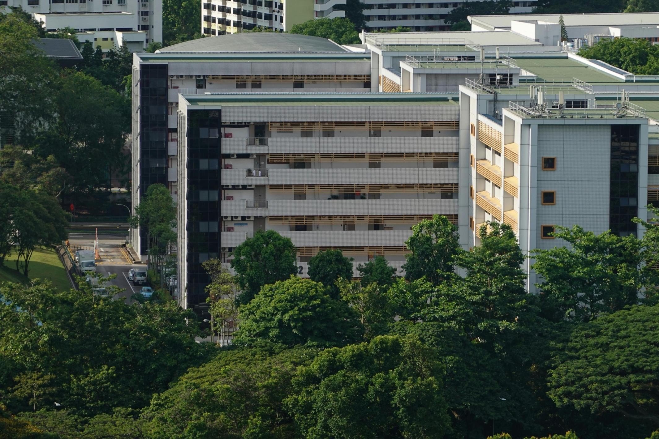Nestled among lush green trees, a modern institutional building features a white facade with horizontal louvers and dark glass accents. The multi-story complex has a flat roof and geometric design, standing in contrast to the glimpse of taller residential blocks in the distance.