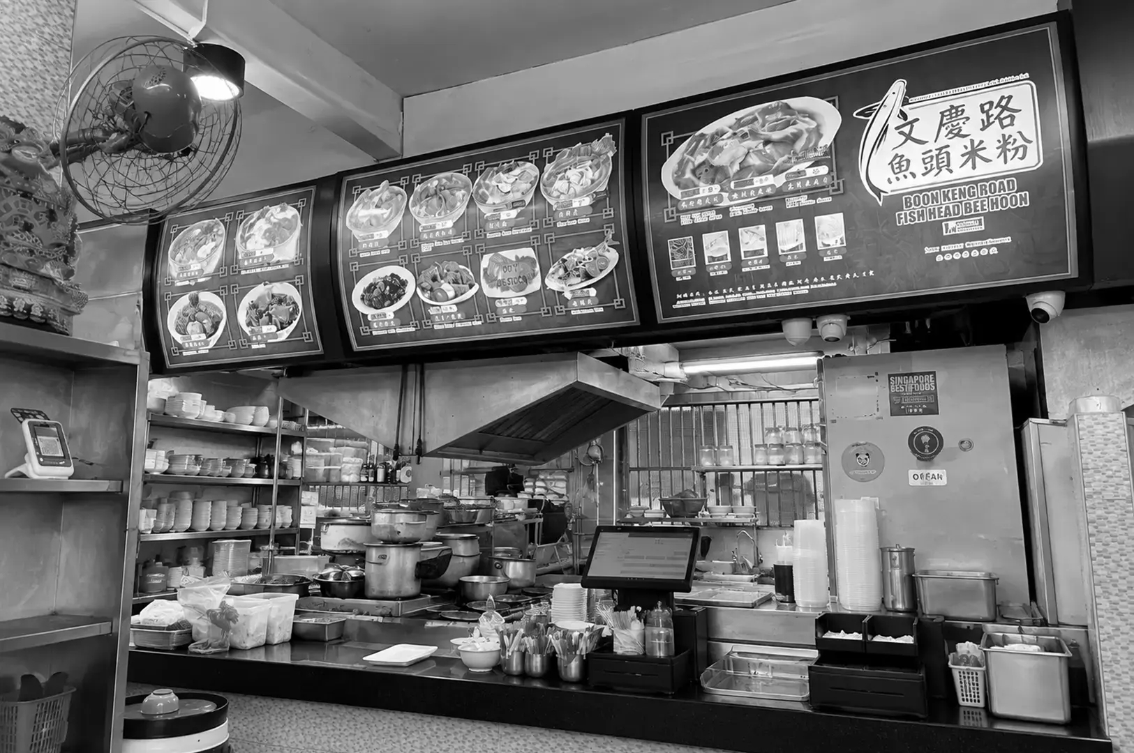 A traditional hawker stall counter displaying food preparation equipment, ingredients, utensils, and overhead menu boards.