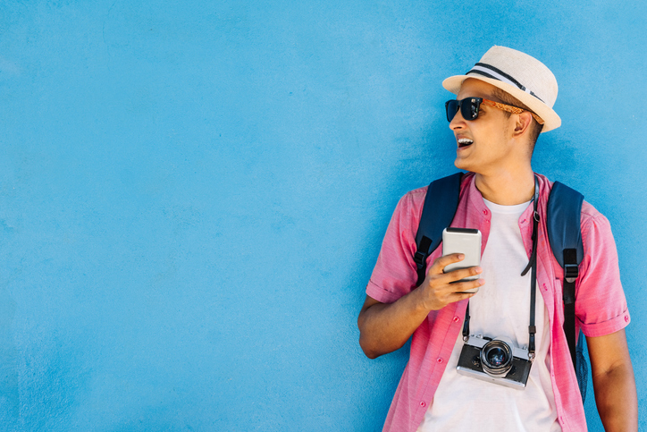 Happy young man in a straw hat with a camera around his neck leaning against a blue wall.  