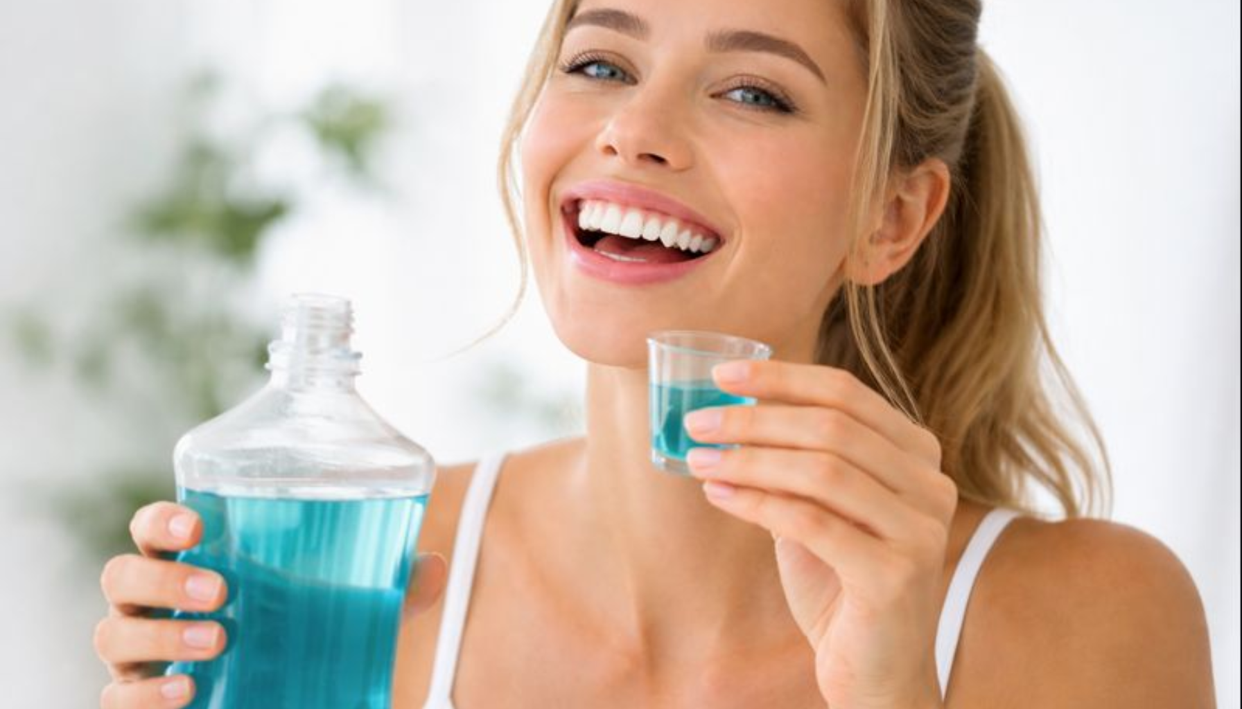 Munnskyll: Når Det Er Nyttig og Når Du Ikke Trenger Det 11 En ung kvinne smiler mens hun holder en flaske og kopp med blå munnskyllevæske i et lyst bad.