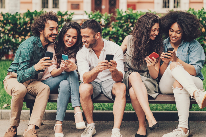 Group of five young adults laughing on a park bench and looking at their phones. 