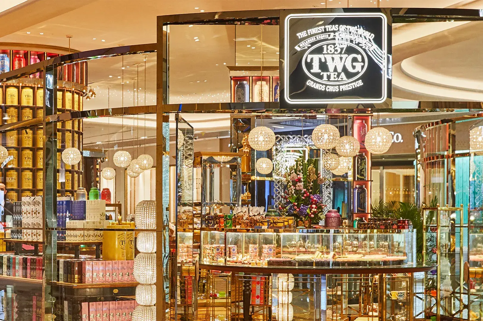 Storefront of a TWG Tea salon featuring brass-framed glass display cases, yellow tea tins, and illuminated crystal pendant globe lights.