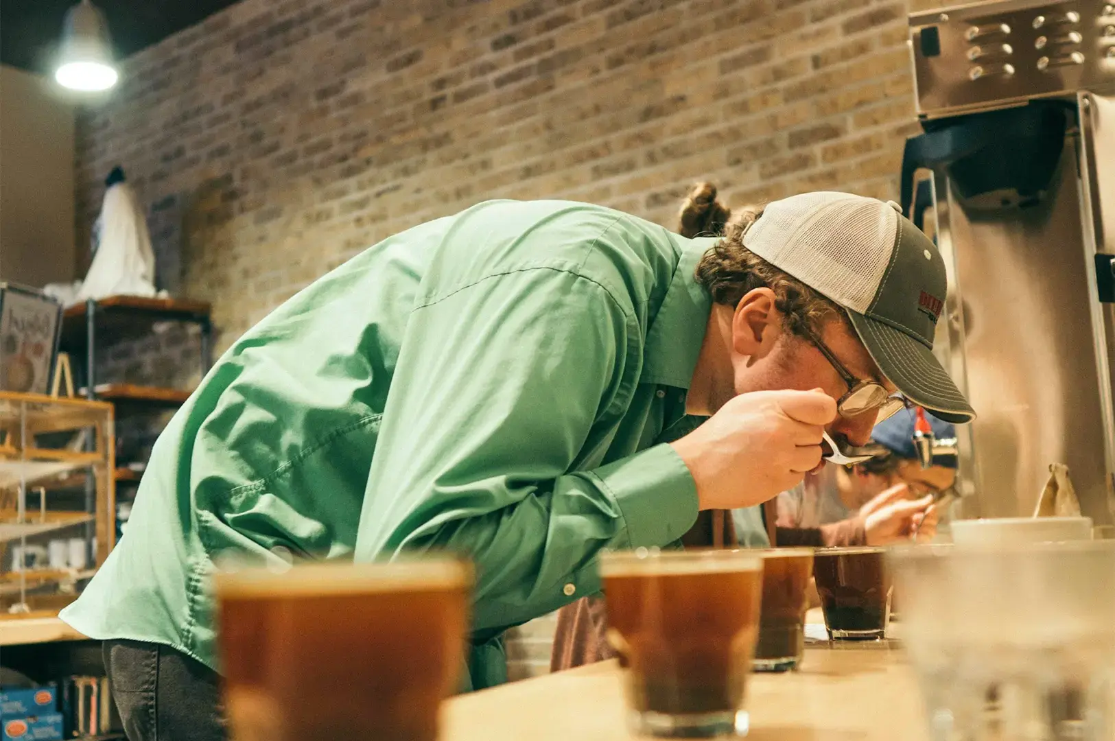 A bartender tasting a freshly brewed coffee under a coffee machine with a line of coffee cups lined at the side.