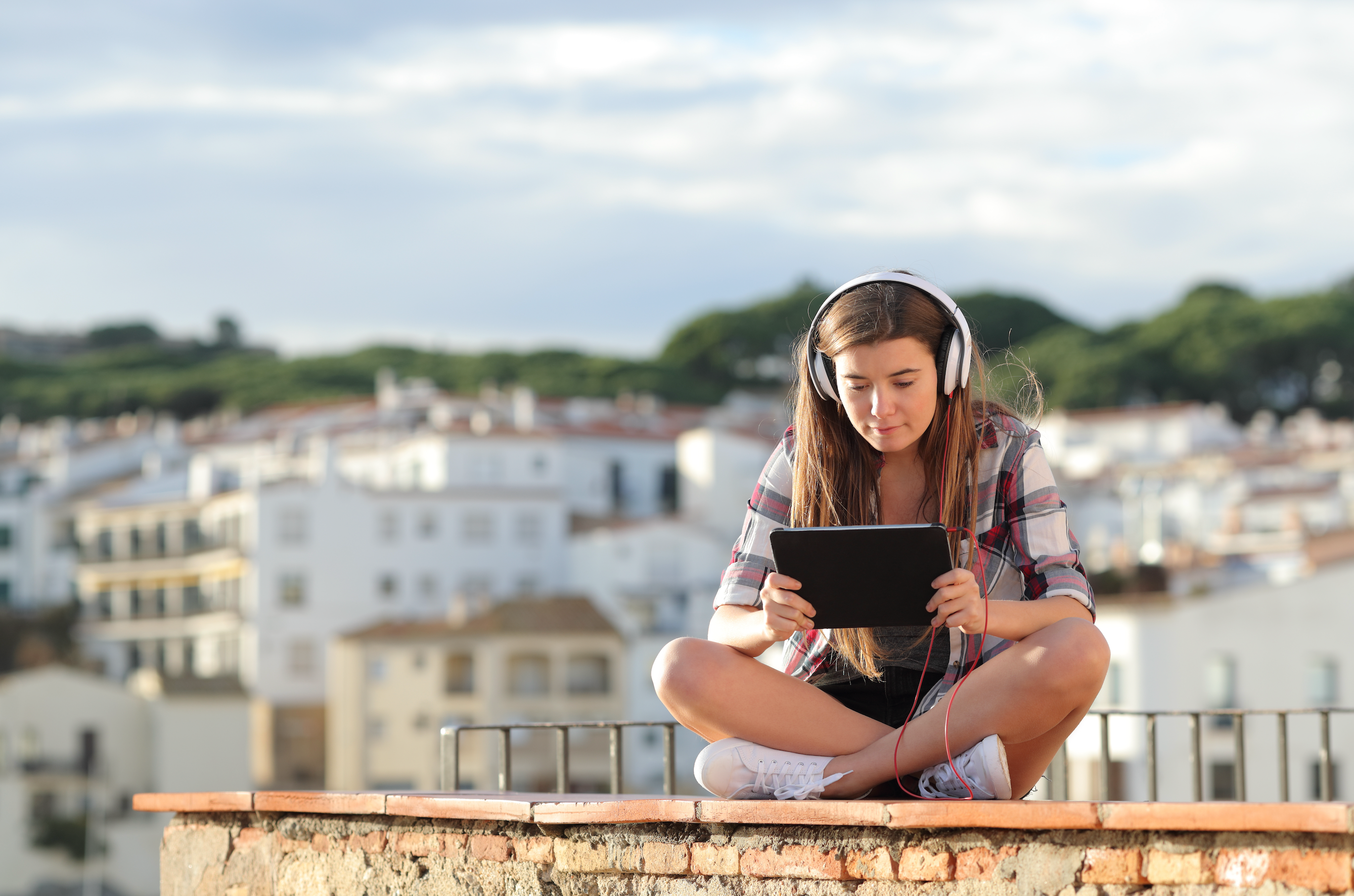 a woman with headphones on watches her tablet while sitting on a brick wall