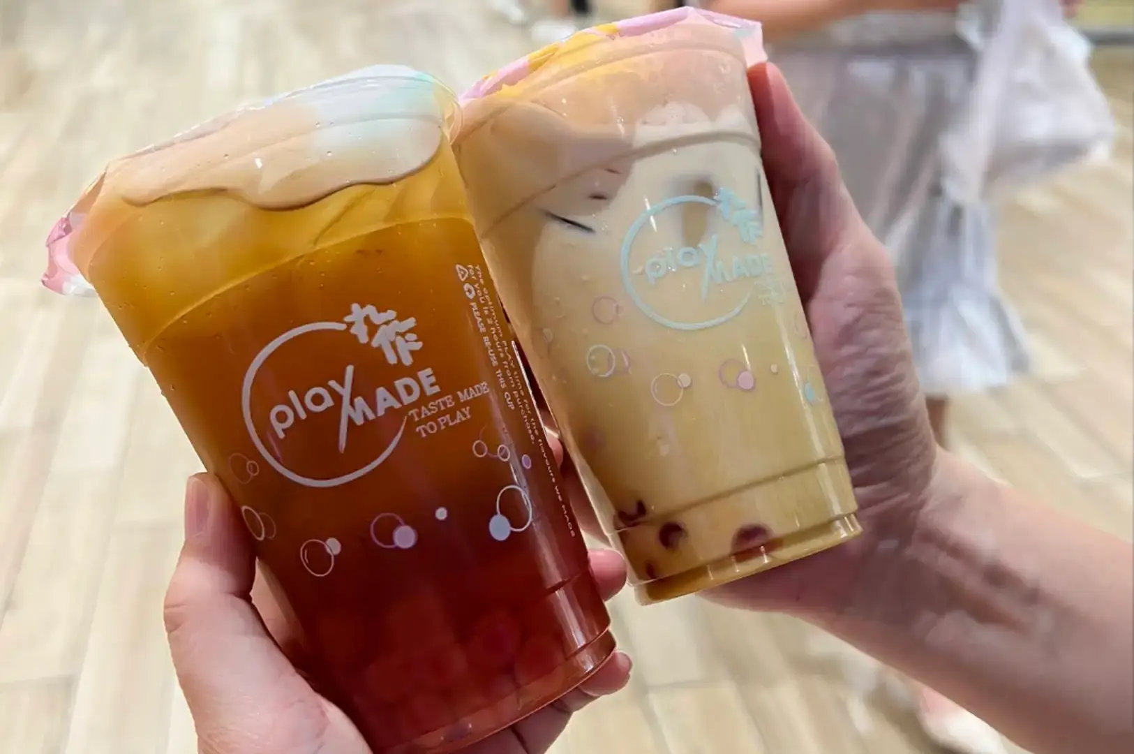 A close-up shot of a hand holding two PlayMade iced bubble teas in clear plastic cups displaying the white logo, one filled with amber iced tea and the other with creamy milk tea, both layered over PlayMade's signature pinkish handmade tapioca pearls at the bottom.