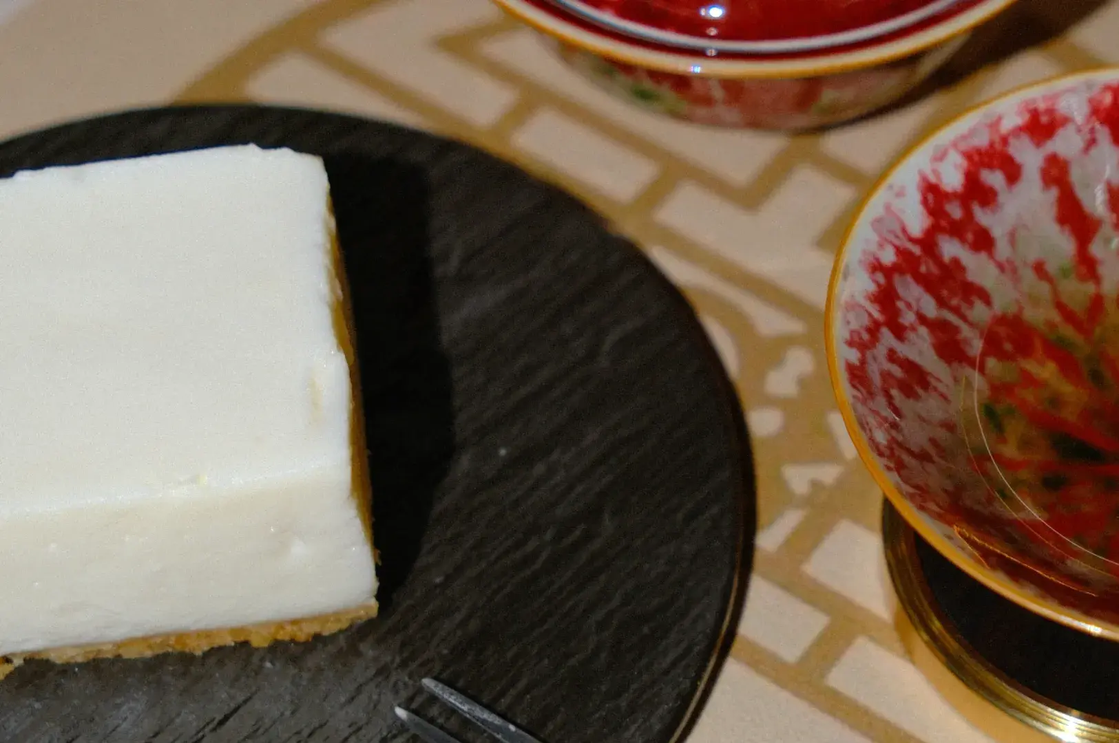 A slice of cheesecake on a black slate plate, accompanied by two red and white decorative bowls. The setting is elegant and inviting.