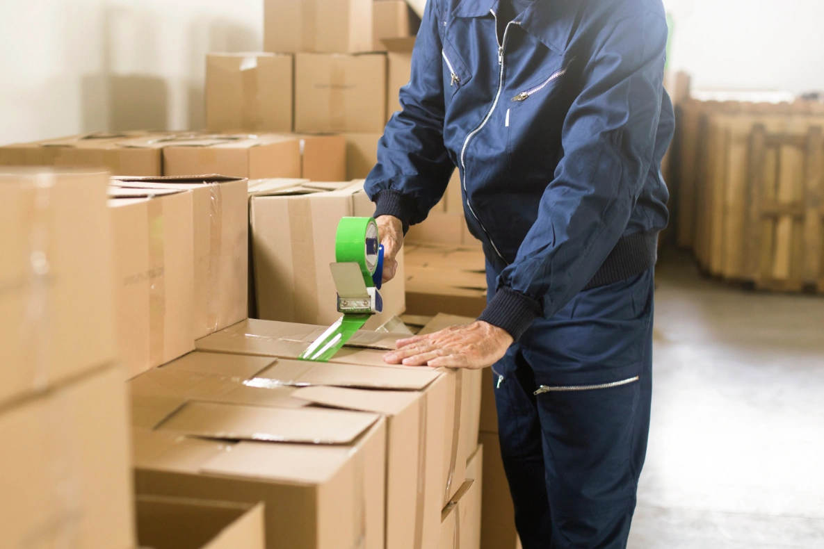 A person in a blue uniform seals a cardboard box with green packing tape in a warehouse full of boxes.