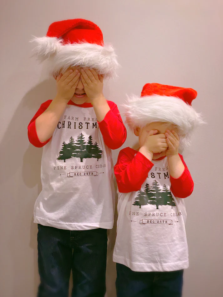 Two boys wearing matching Christmas outfits—(raglan shirts), pants, and Christmas hats—covering their face