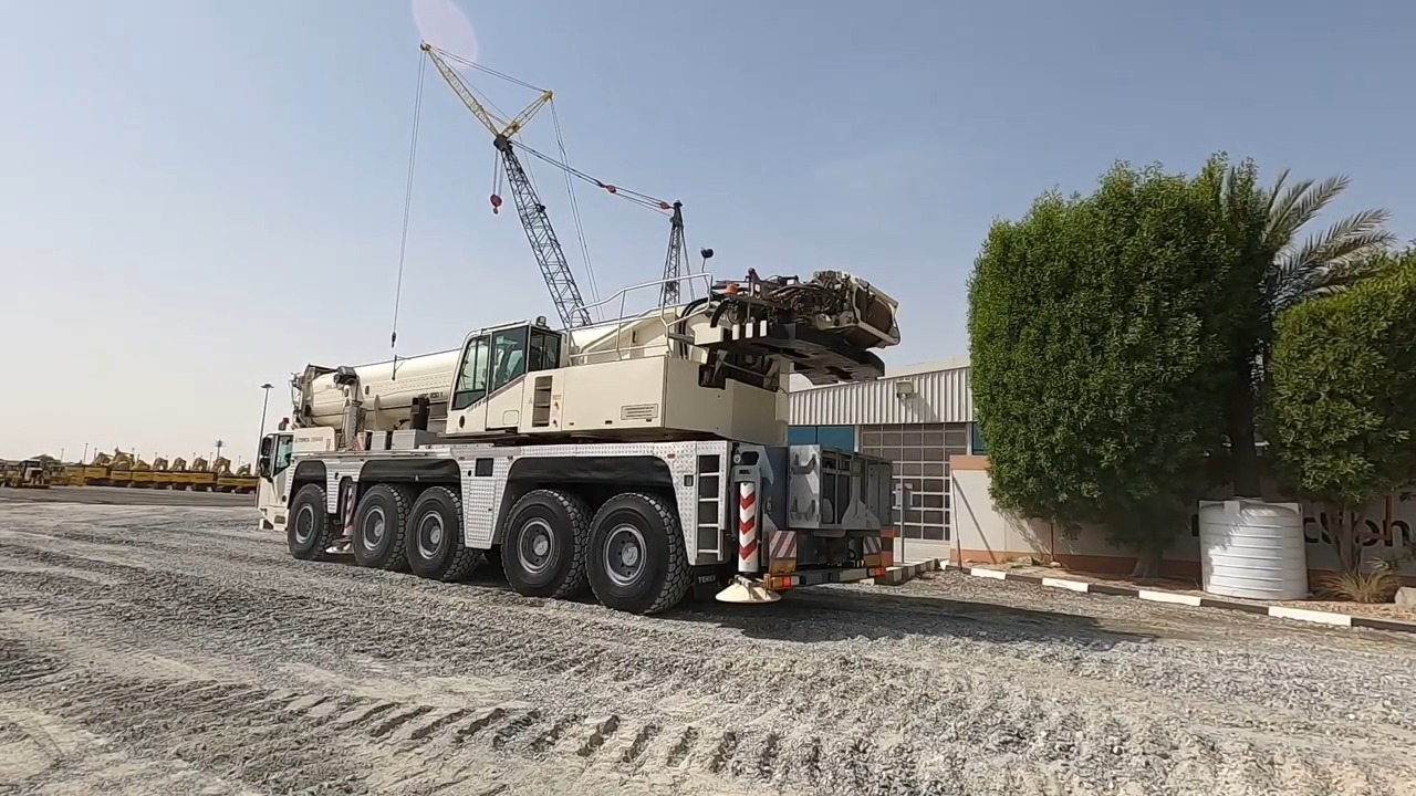 Large multi-wheeled mobile crane parked on a gravel lot in an industrial area, with its boom extended upward near other construction equipment and greenery