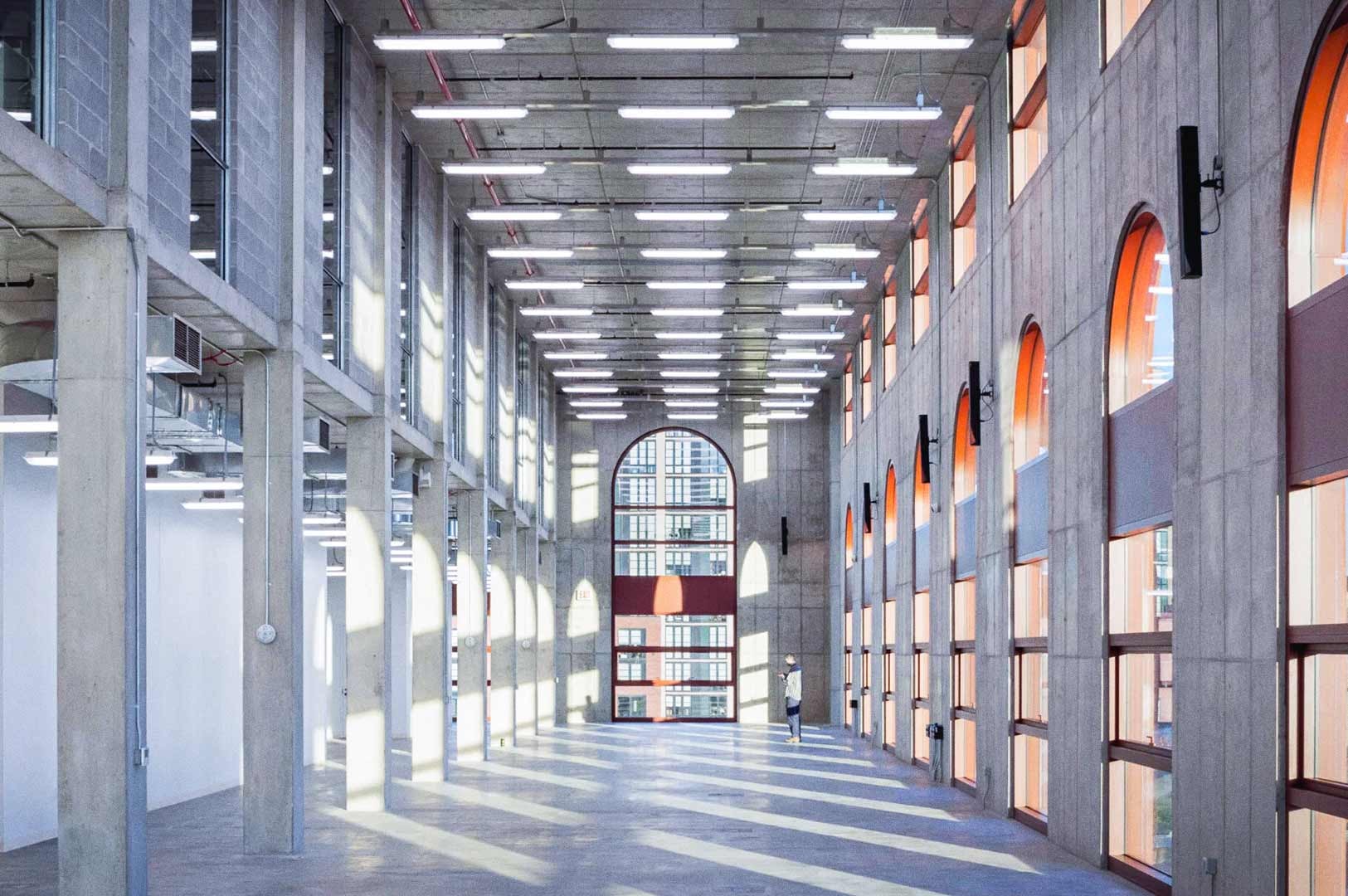 Spacious interior of a renovated industrial building with tall concrete columns, high ceilings, and rows of large arched windows letting in bright sunlight. A single person stands near the far end of the open hall.