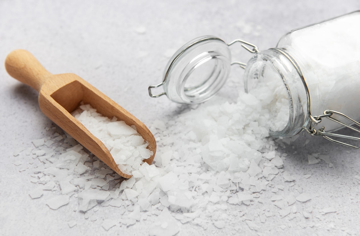 Epsom salt flakes spilled from glass jar with wooden scoop on light surface, natural magnesium sulfate bath soak ingredient