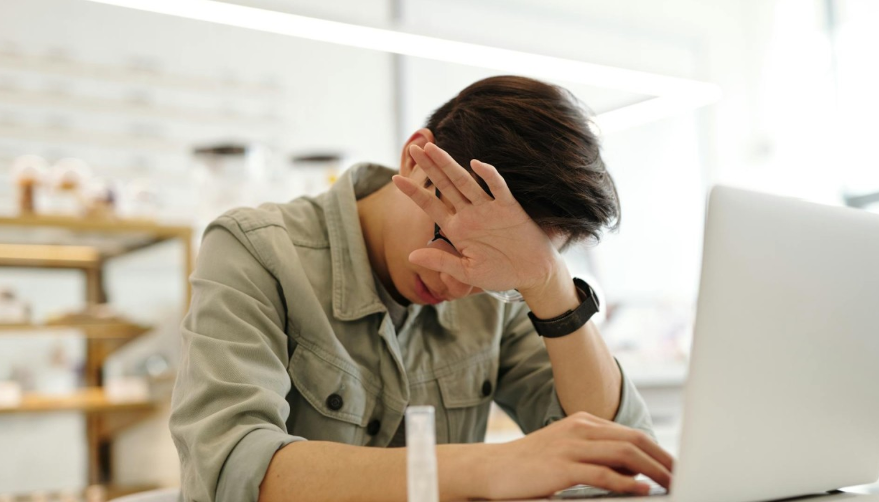 Ung mann foran bærbar PC dekker ansiktet med hånden, ser utmattet eller frustrert, muligens stresset eller sliten på jobb eller skole.