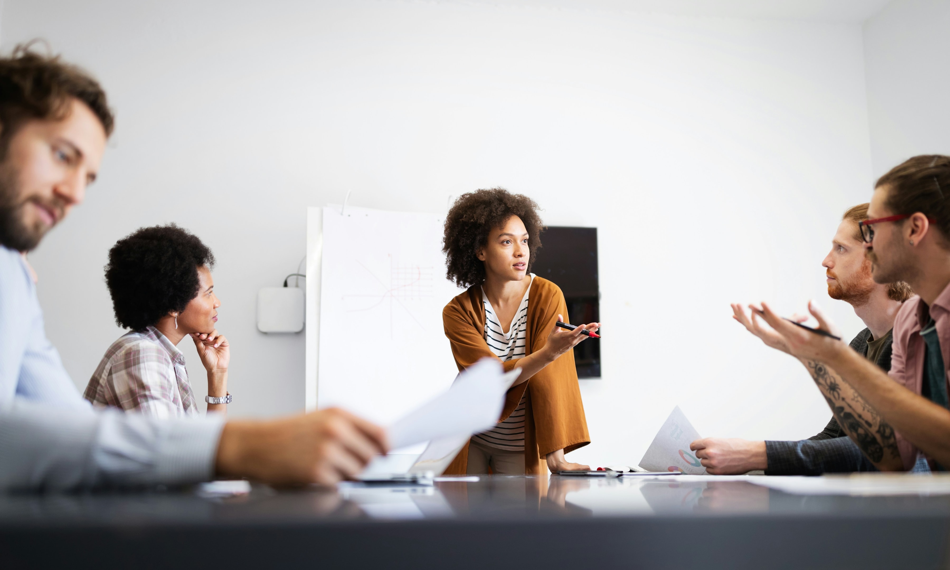 Woman in brown jacket talking to team who sit around table
