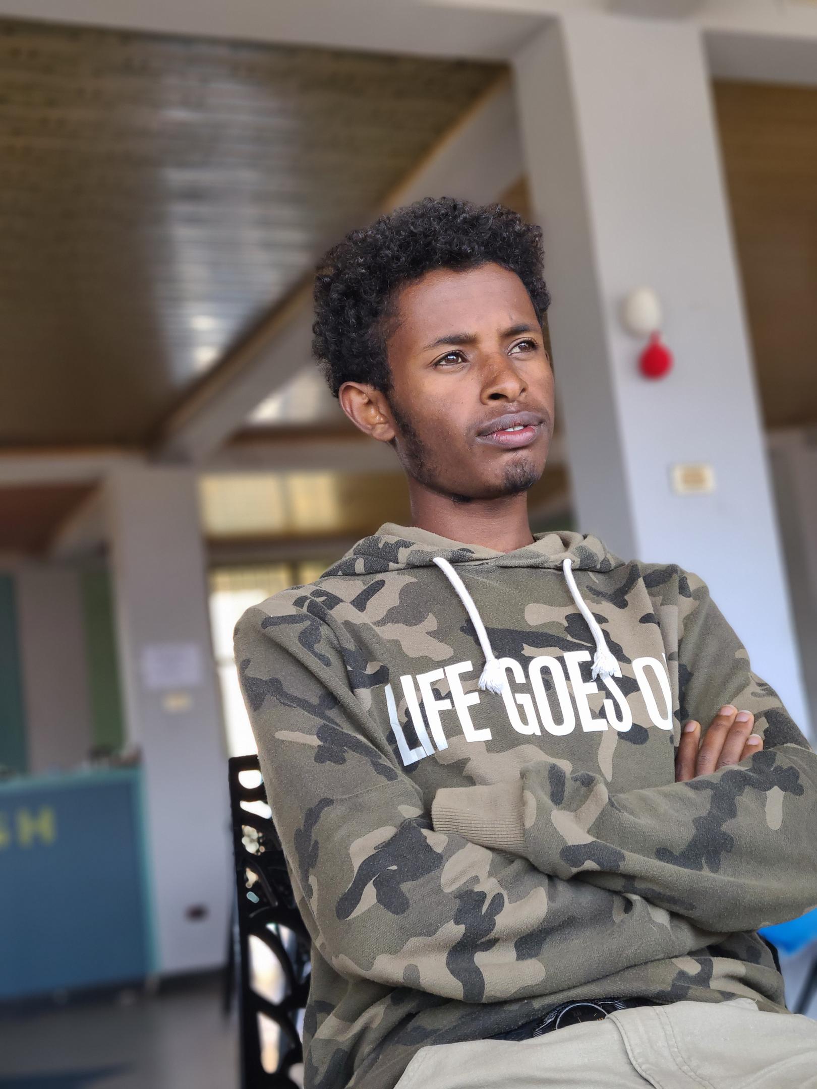 A young man sits with arms crossed, gazing thoughtfully into the distance, wearing a camouflage hoodie printed with the phrase "Life Goes On," set against the blurred interior of a public building.