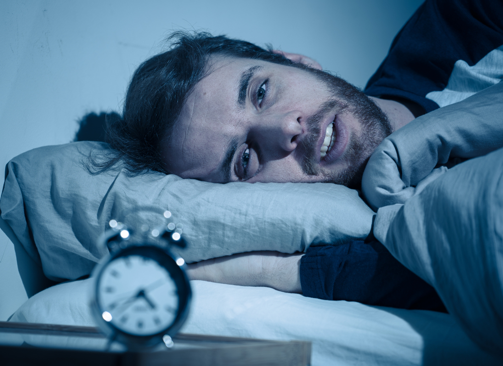 Man looking exhausted with a clock at 4:37AM suffering from insomnia. Junk food causes anxiety and sleep problems. 