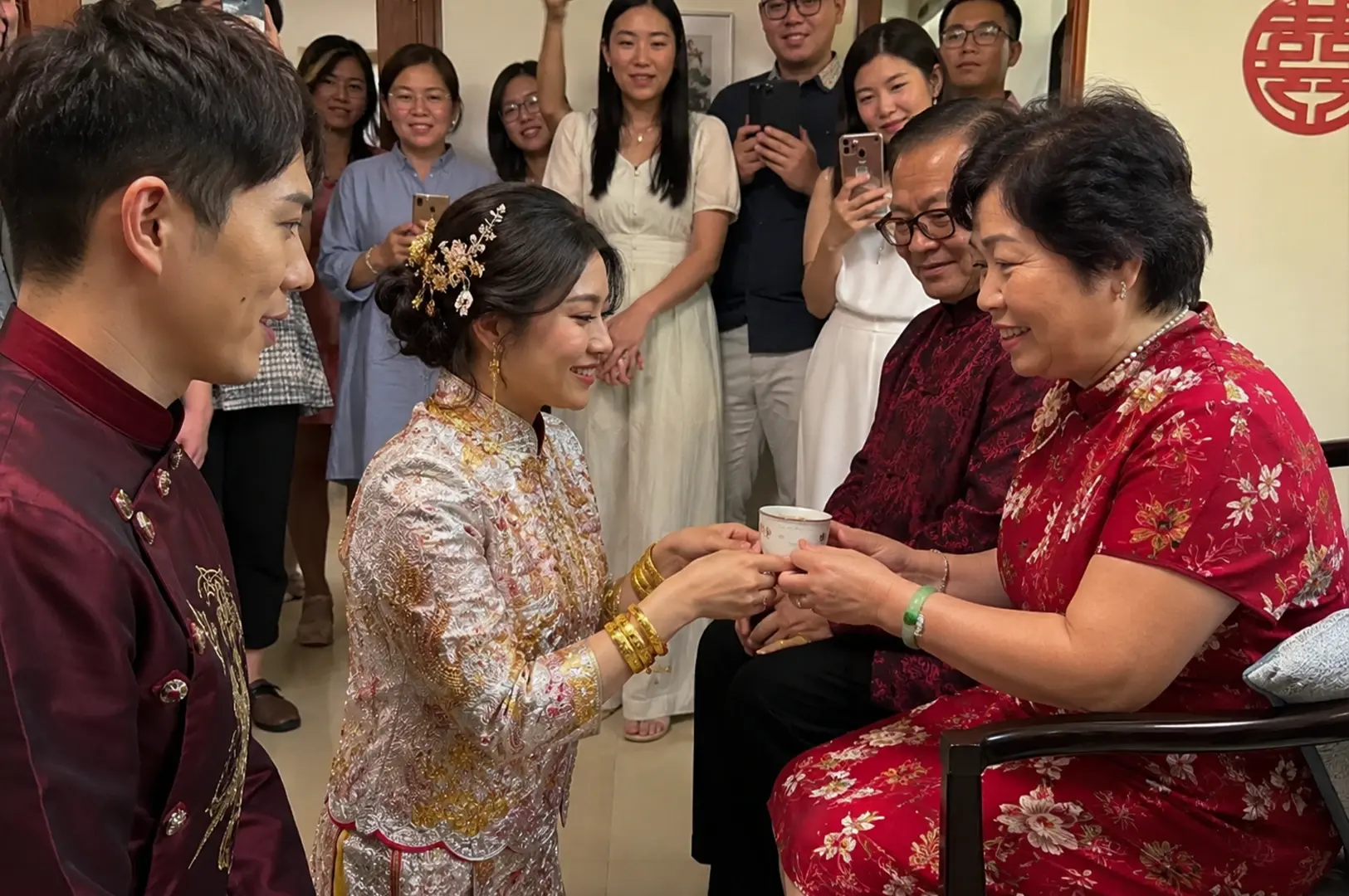 A bride in a white and gold embroidered Qun Kwa dress knees down, smiling as she presents a small white tea cup to an older woman wearing a red floral cheongsam dress. The bride wears heavy gold bangles on both wrists and an intricate gold floral hairpin in her updo, while the groom, dressed in a dark maroon embroidered tunic, stands close by her side. The older woman sits next to a man in glasses and a dark red shirt, accepting the tea with a smile. Behind them, a crowd of smiling family members stands in a doorway, holding up smartphones to capture the traditional ceremony beneath a red "Double Happiness" sign on the wall.