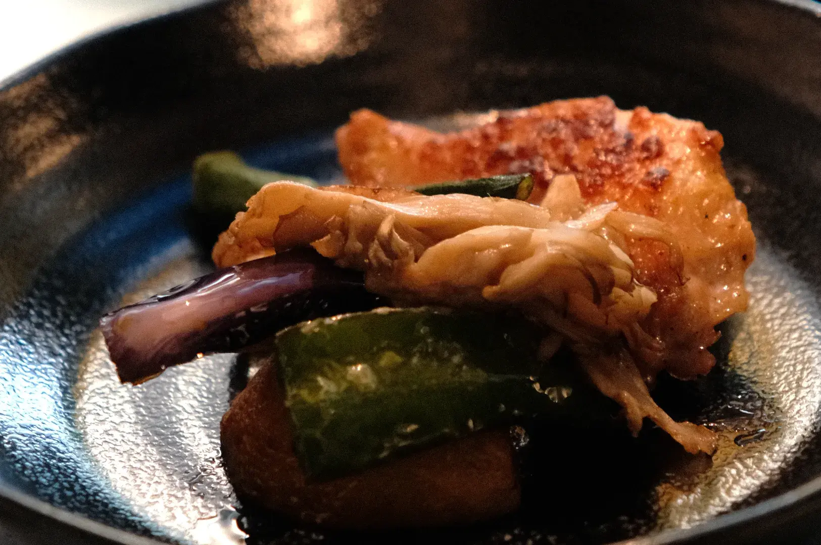 A piece of cooked chicken with green peppers and eggplant on a dark plate.