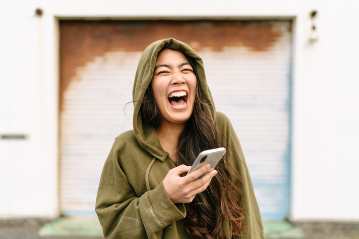 Pretty young woman wearing a green hoodie laughing hysterically at something she read on her cell.