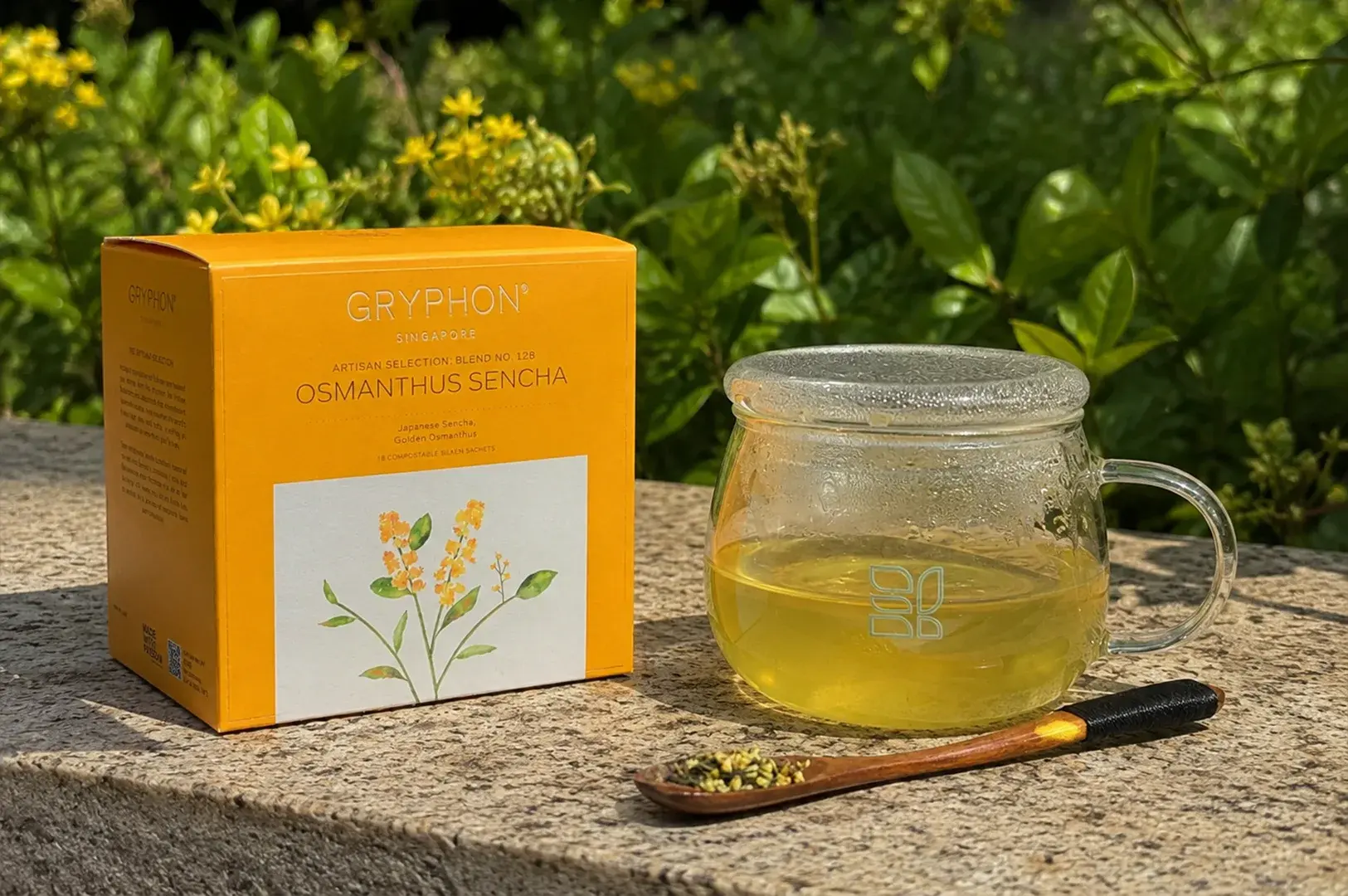 An eye-catching, ochre-yellow box of Gryphon "Osmanthus Sencha" tea rests on a textured stone ledger or retaining wall, backlit by a sunlit garden of vibrant green bushes and clusters of yellow flowers. A clear glass mug filled with a pale, yellow-green steeped sencha tea sits beside the box, showing a modern, minimalist square logo etched into the glass. A slim wooden spoon holding a small pile of dried green tea leaves and tiny yellow osmanthus flowers lies in the foreground on the stone surface.