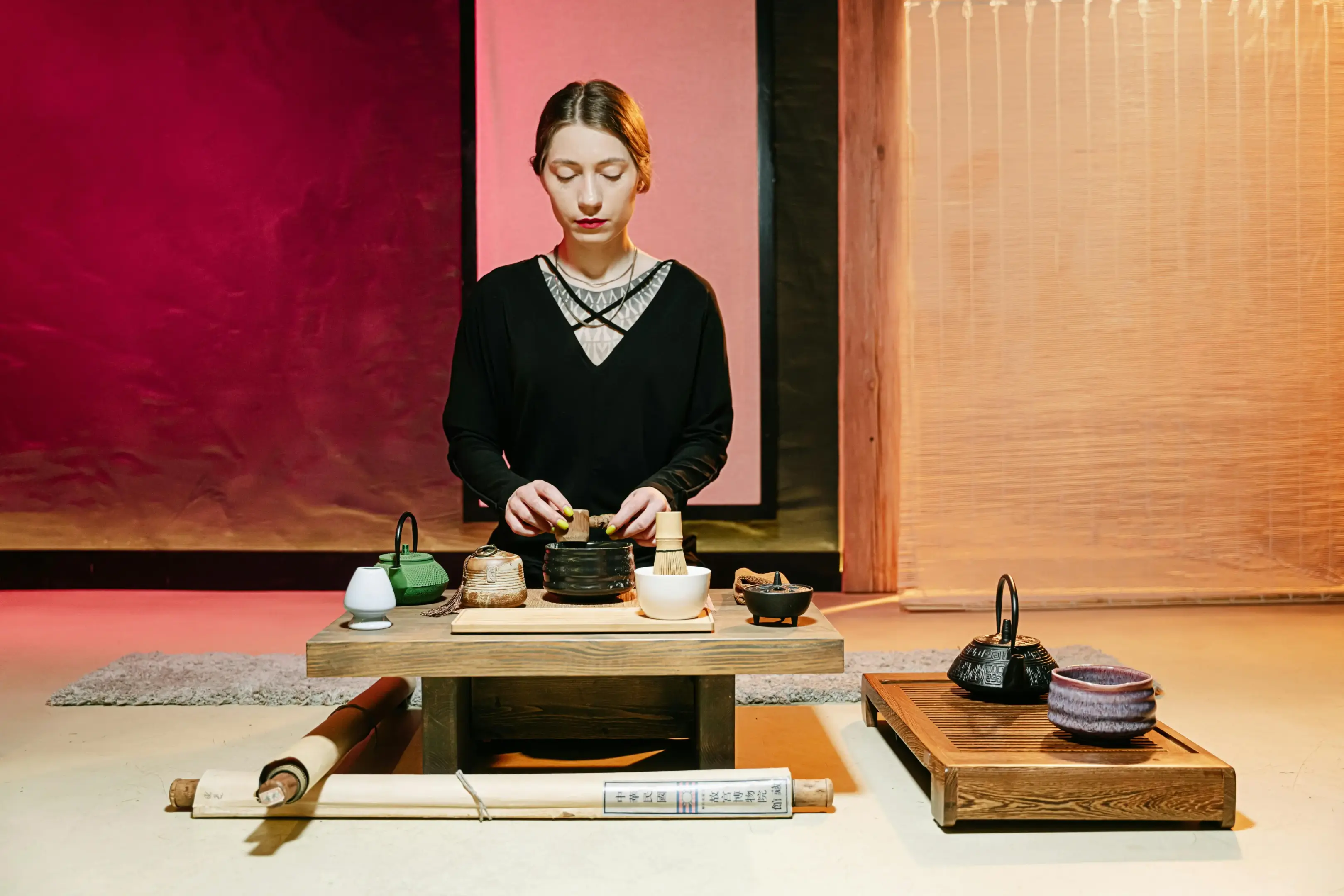 A woman dressed in black sits calmly behind a low wooden table, meticulously preparing tea with traditional Japanese matcha tools. The serene setting features vibrant red and soft pink backdrops, complemented by various teapots and scrolls laid out on the floor.