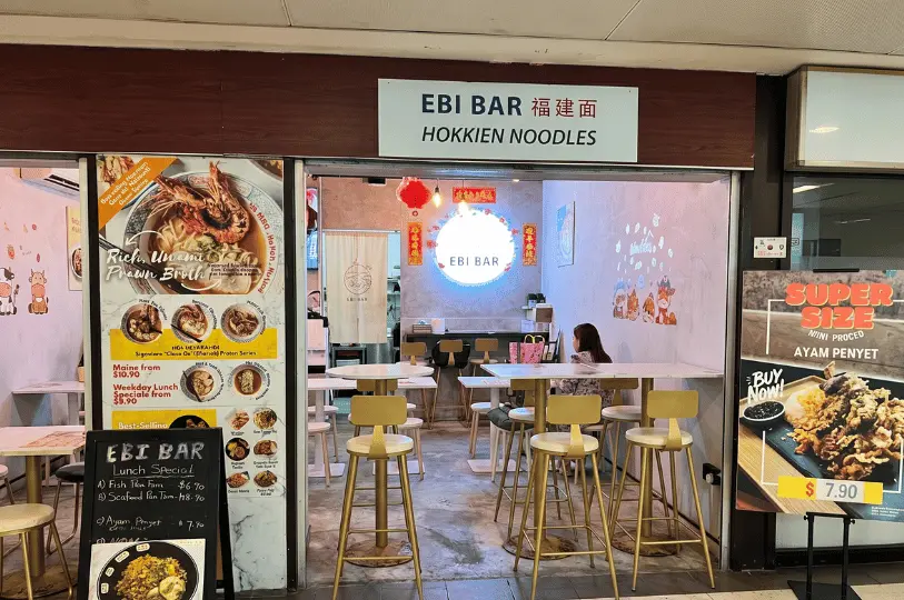 The modern, brightly lit storefront of Ebi Bar Hokkien Noodles, showcasing promotional food posters outside and high tables with gold-colored stools inside.