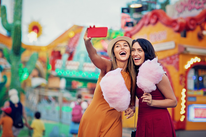 Happy young women holding cotton candy and taking a selfie at an outdoor fair.