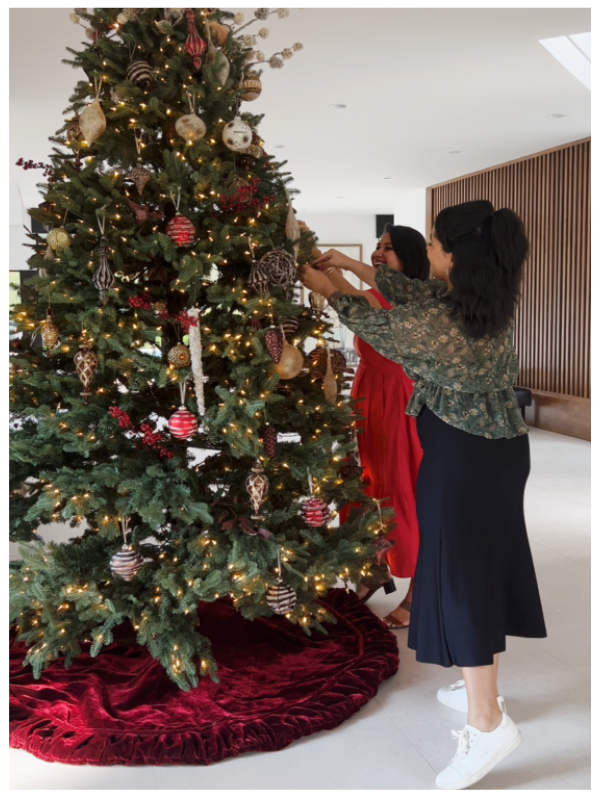 Two women decorate a tall Christmas tree with red and gold ornaments and warm string lights
