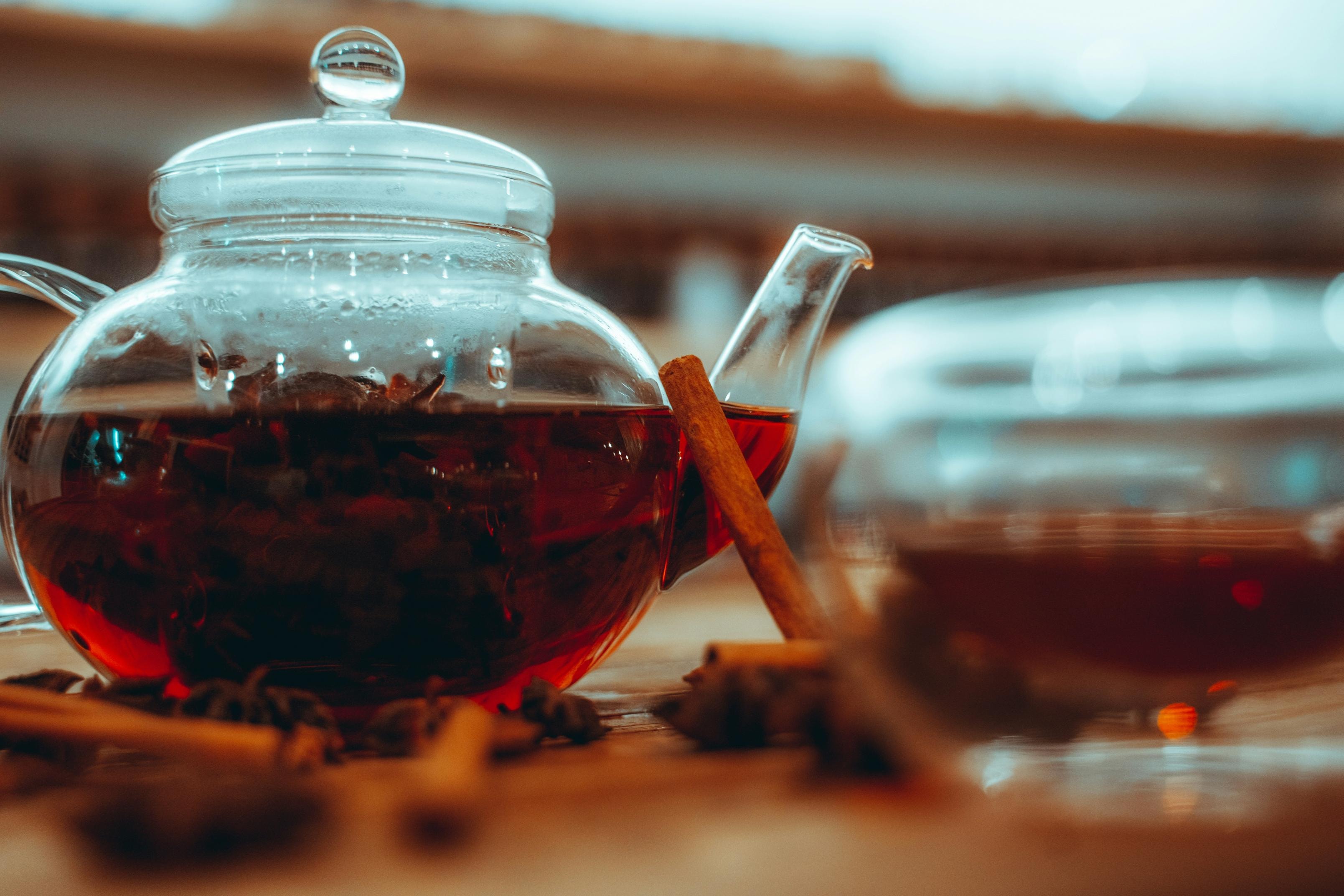 A clear glass teapot filled with a dark, reddish-brown tea sits on a wooden surface surrounded by aromatic cinnamon sticks and star anise. The soft, blurred background emphasizes the warm colors of the tea and spices, creating an inviting and cozy atmosphere.