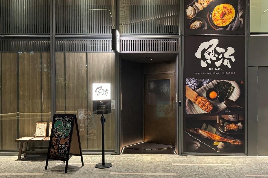  The image displays the modern and sleek exterior of Uoharu, a Japanese restaurant featuring a dark, recessed entryway and large window panels adorned with vibrant food photography. A chalkboard menu stand and a glowing logo sign greet customers at the entrance, suggesting a refined yet welcoming atmosphere for diners in Singapore.